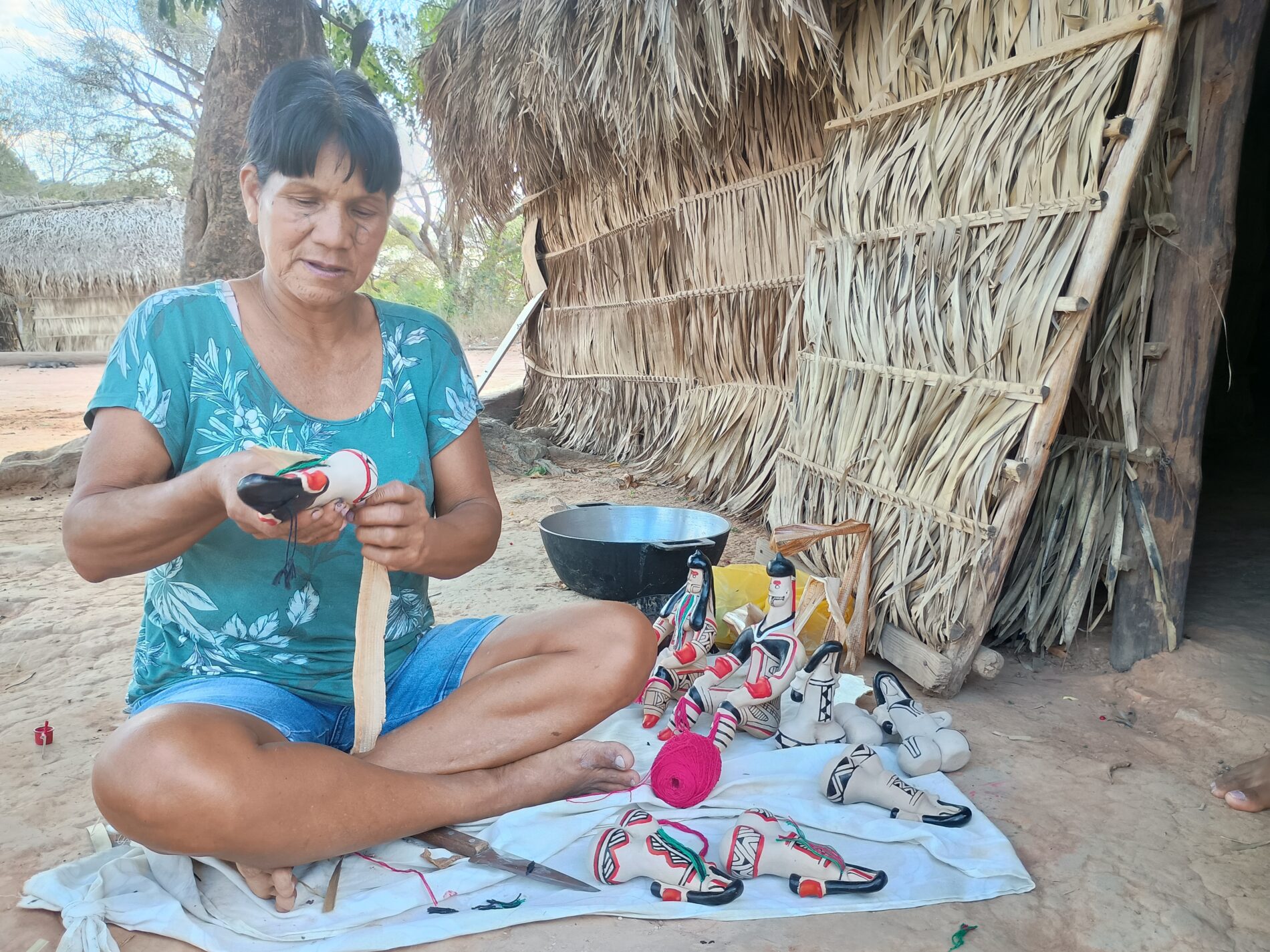 Artesã Karajá segura Ritxoko, conhecida como "Bonecas Karajá". Ela está sentada em um bano, de pernas cruzadas e usa um vestido azul estampado.